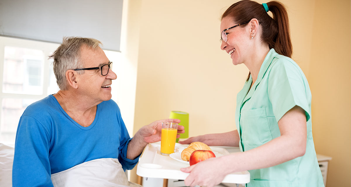 Happy Residents Smliling nurse wearing green serving a tray of food to happy patient wearing a blue shirt