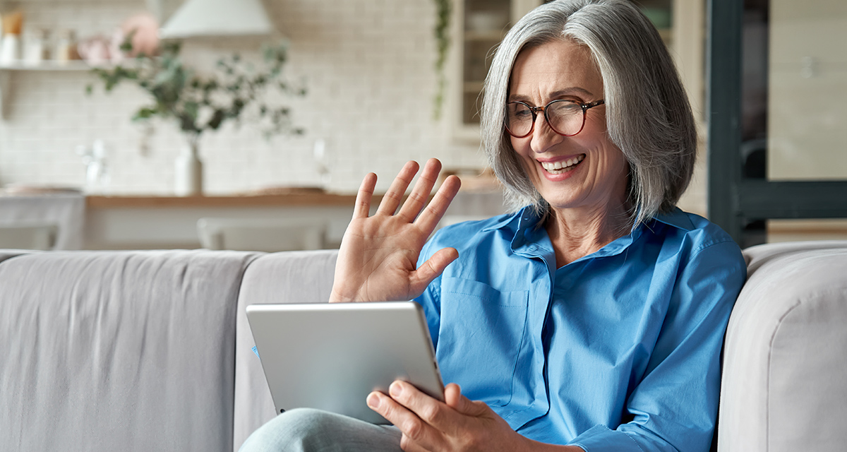 Older Adults Older grey haired woman in blue shirt sitting on couch smiling while holding a tablet