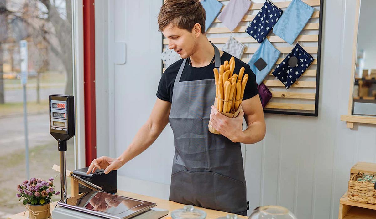 Support Quick Service Options & Self-Service Kiosks Young man waring apron holding breadsticks using a point of sale system