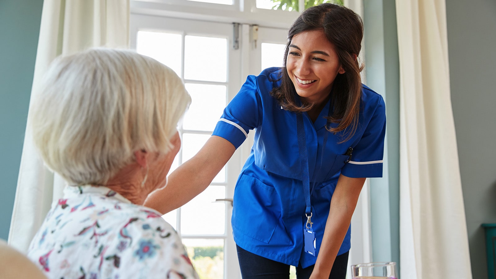 Resident Care Nurse wearing blue smiling at elderly resident after delivering meal
