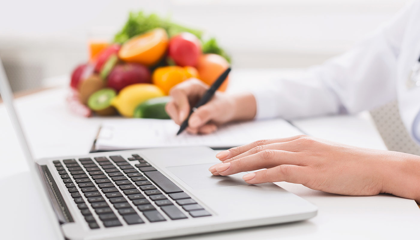 Nutritionist Nutritionsit using laptop and writing down notes with stack of fruit in the background