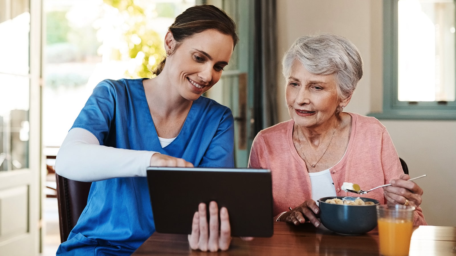 Easy to Use Nurse in blue smock showing elderly woman wearing pink something on a tablet while she eats breakfast